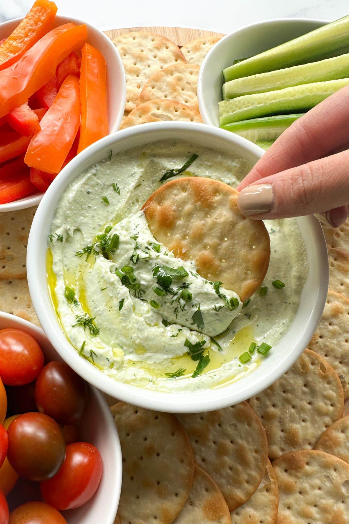 Hand dipping cracker into cottage cheese dip with veggie dippers in background.