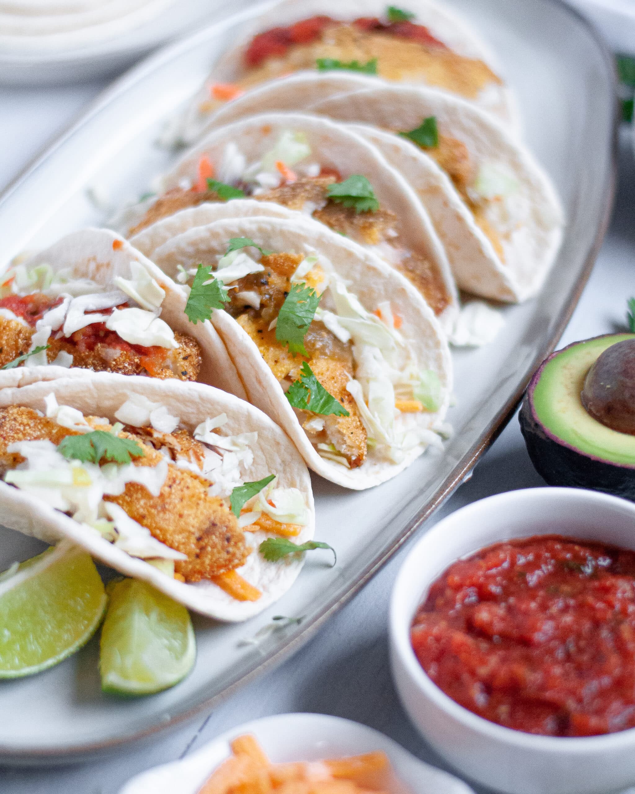 Cornmeal crusted fish tacos on a platter with lime wedges, salsa, and avocado in the background.