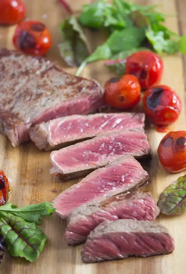 Sliced stead with roasted tomatoes on a wooden cutting board.