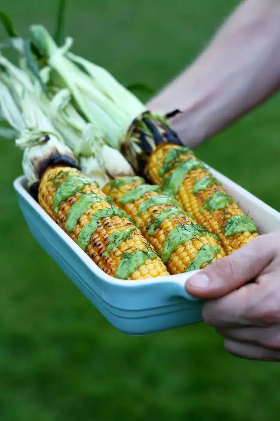 Hands holding baking dish filled with grilled corn on the cob drizzled with avocado dill dressing.