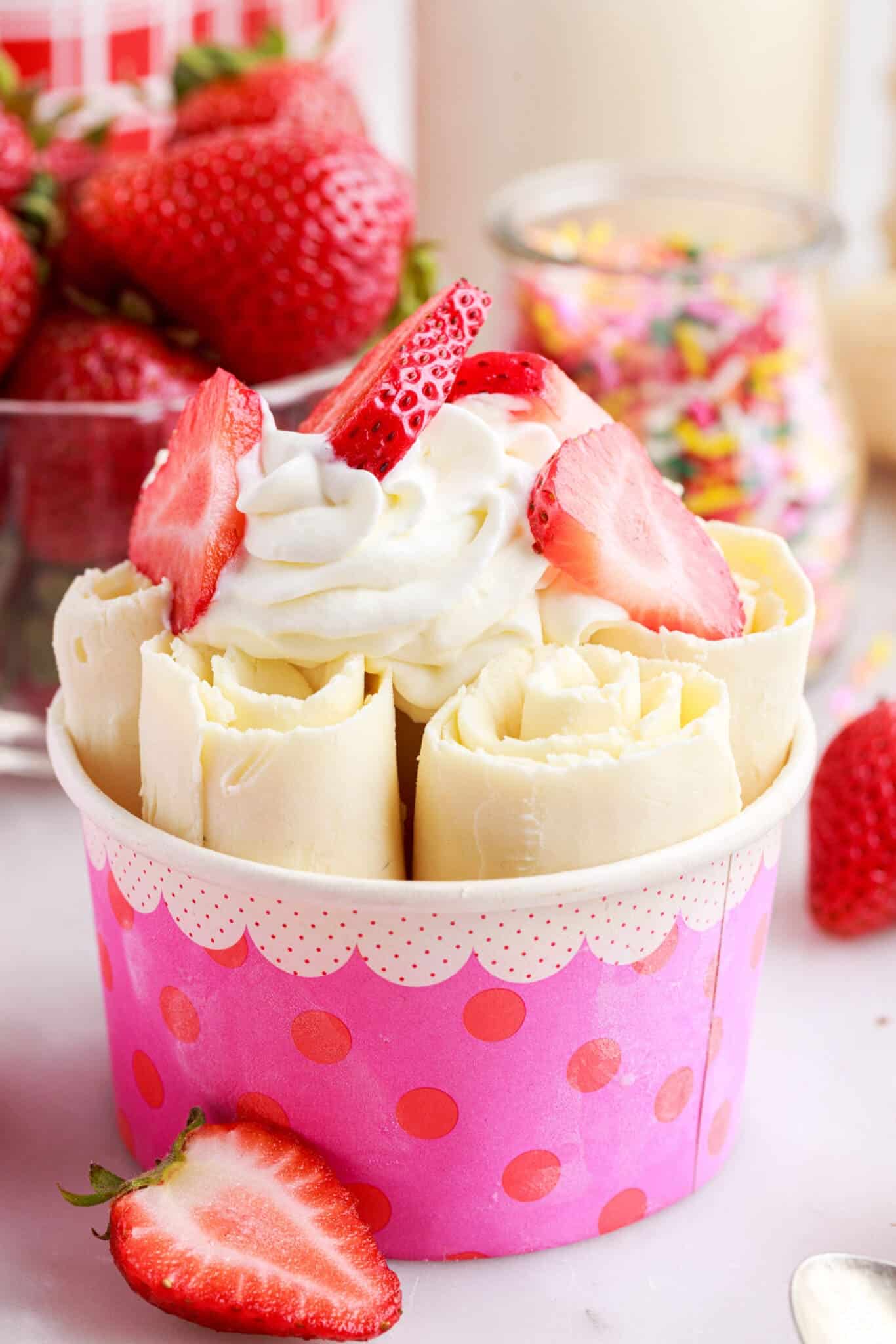 Rolled ice cream with whipped cream and sliced strawberries on top and more strawberries and sprinkles in the background.