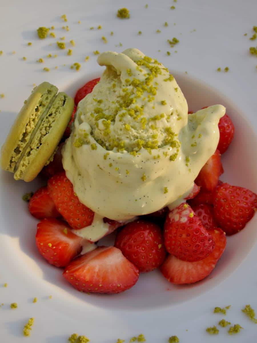 White bowl full of wasabi ice cream with crushed pistachio, macaron, and sliced strawberries.