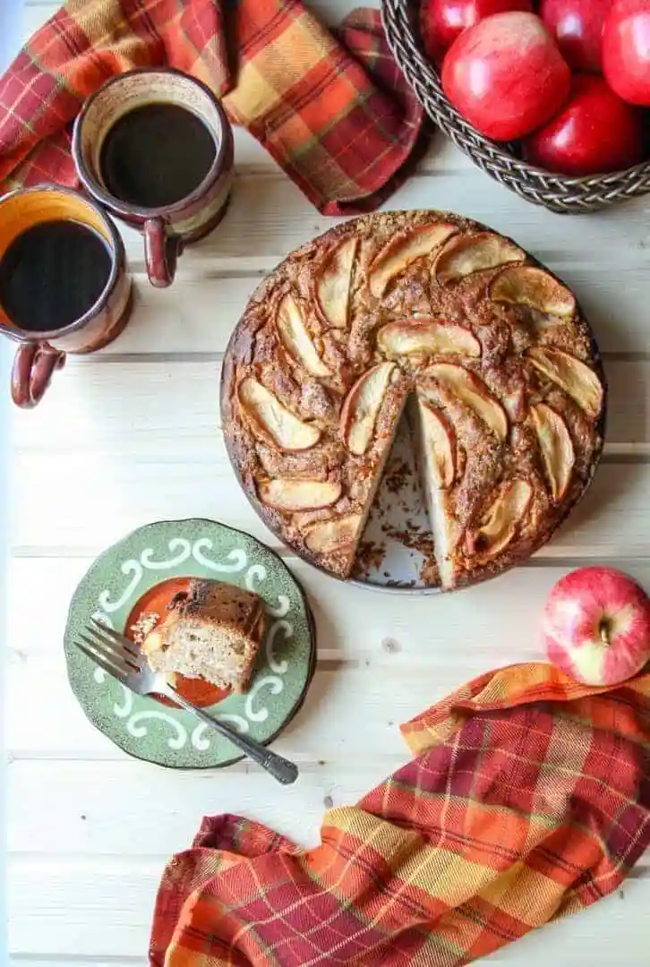 Apple ricotta cake with coffee and basket full of apples in the background.