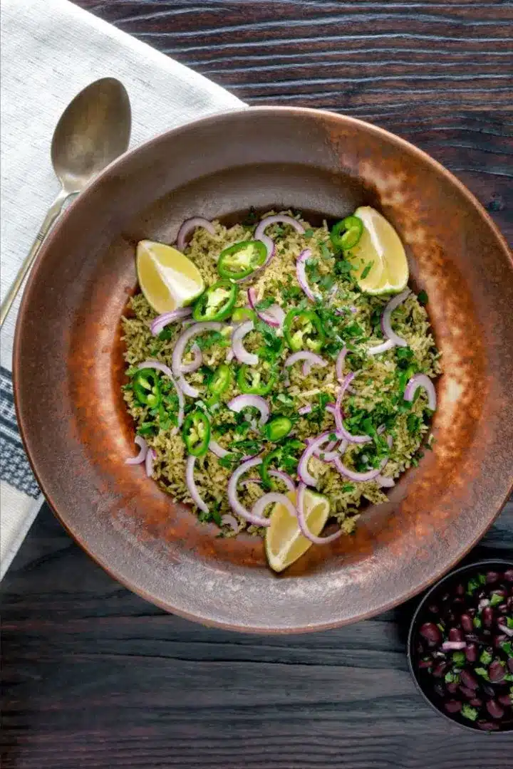 Mexican green rice in a brown bowl with red onion and lime wedges with spoon on the side.