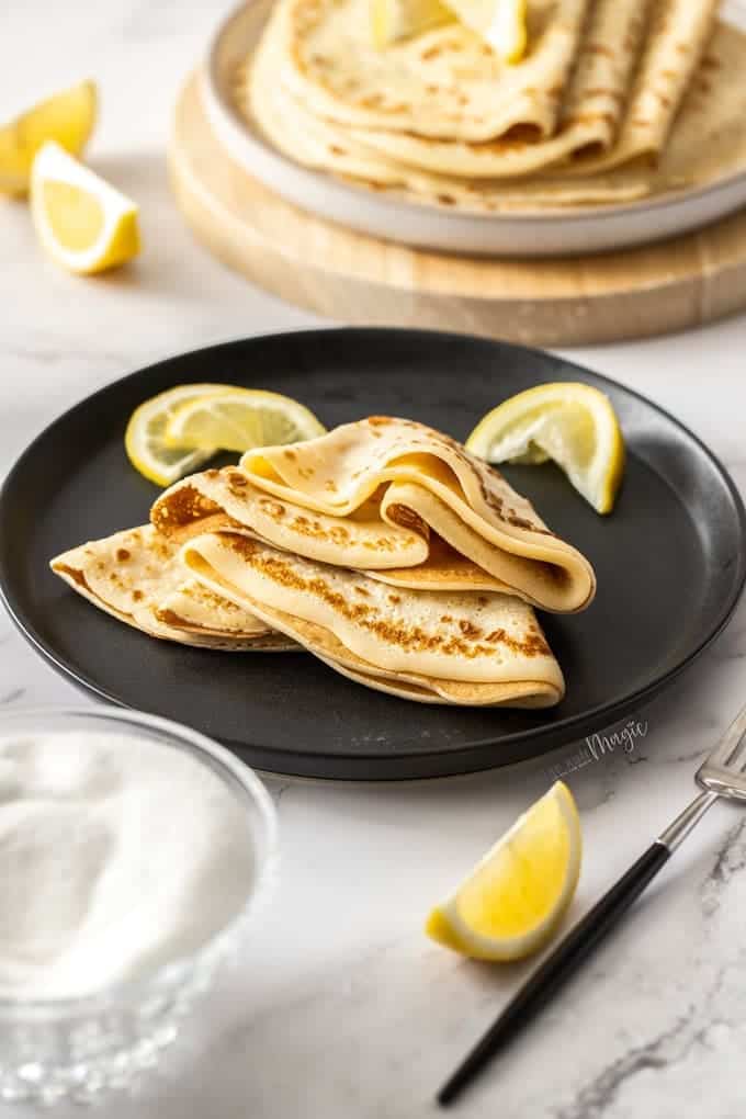 Thin pancakes on a black plate with lemon wedges.