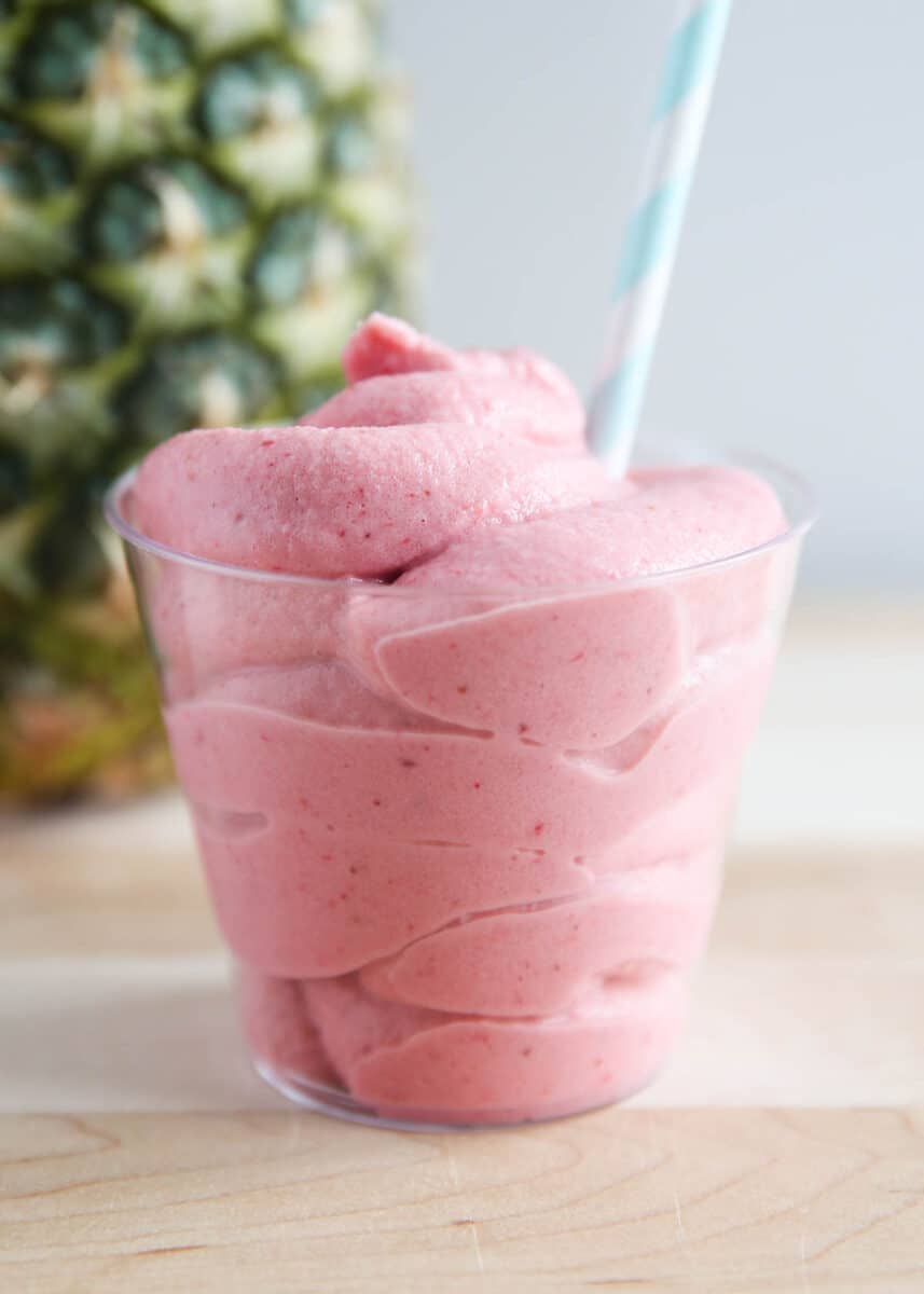 Glass filled with strawberry Dole whip with a striped straw and a pineapple in the background.