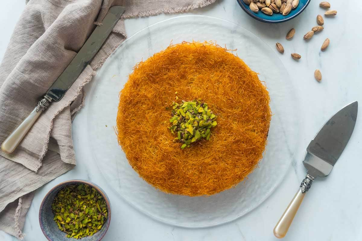 Overhead shot of kunafah dessert with knife and nuts in the background.