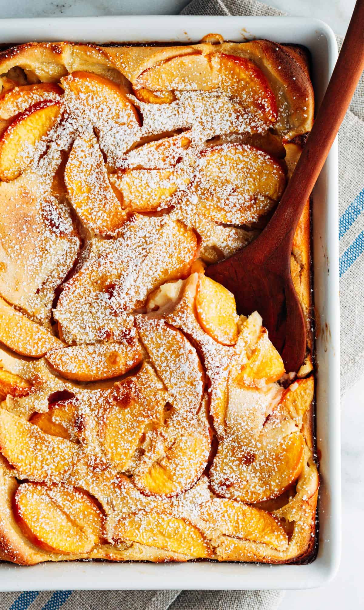 Wooden spoon with a baking dish full of peach clafoutis.