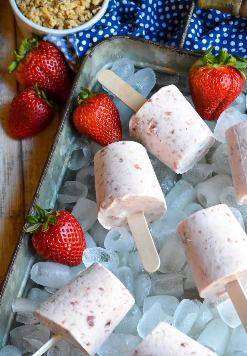 Strawberry creamy ice cream pops on ice with whole strawberries in background.