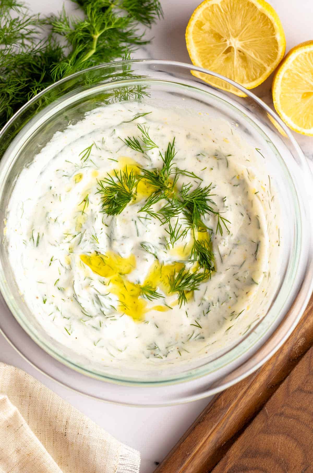 Yogurt dill sauce in a bowl with lemon and herbs surrounding it.