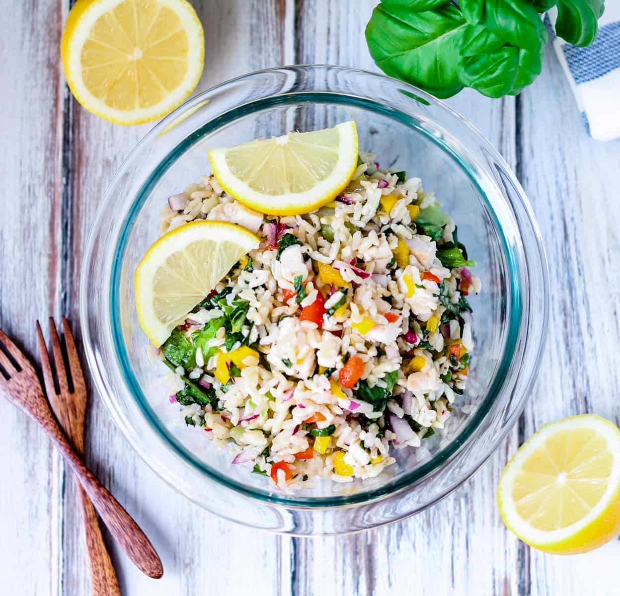 Clear bowl of chicken and rice with fresh basil and lemon with fresh basil and sliced lemon on a white board.