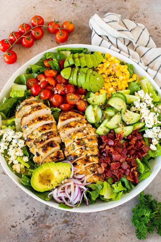 Salad with many ingredient in a large bowl including grilled chicken, cucumbers, corn, avocado, blue cheese, and bacon.