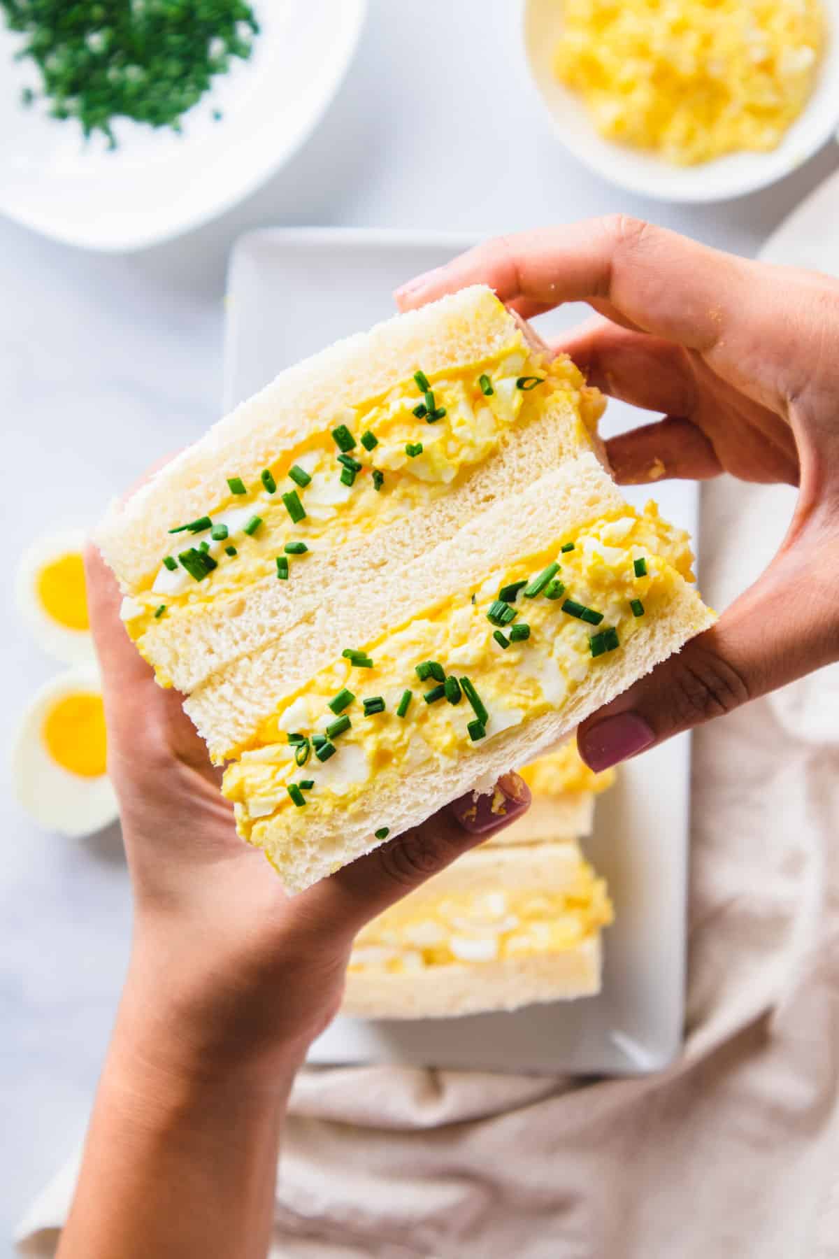 Hands holding cut in half egg salad sandwich.