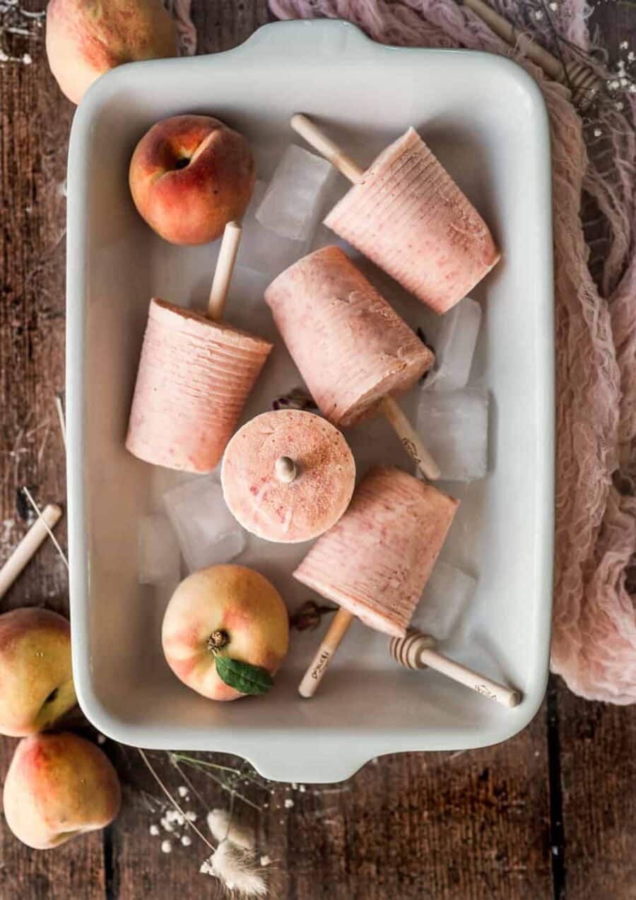 Peach colored popsicles with ice and peaches in a white dish.