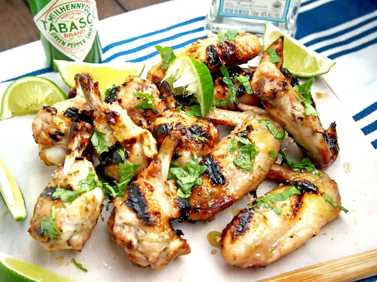 Grilled margarita wings on a platter with limes and hot sauce in the background.