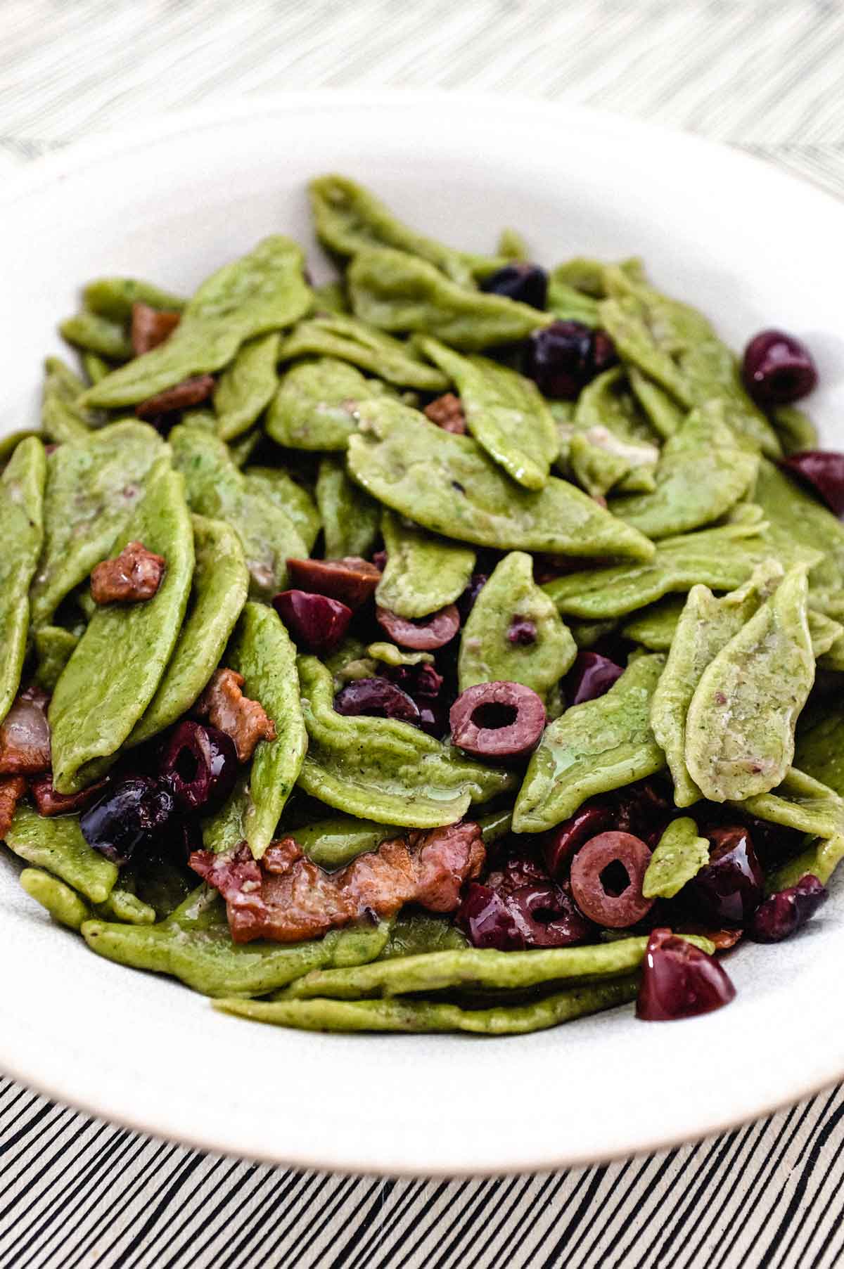 Green pasta with olives in a blue bowl on top a striped surface.