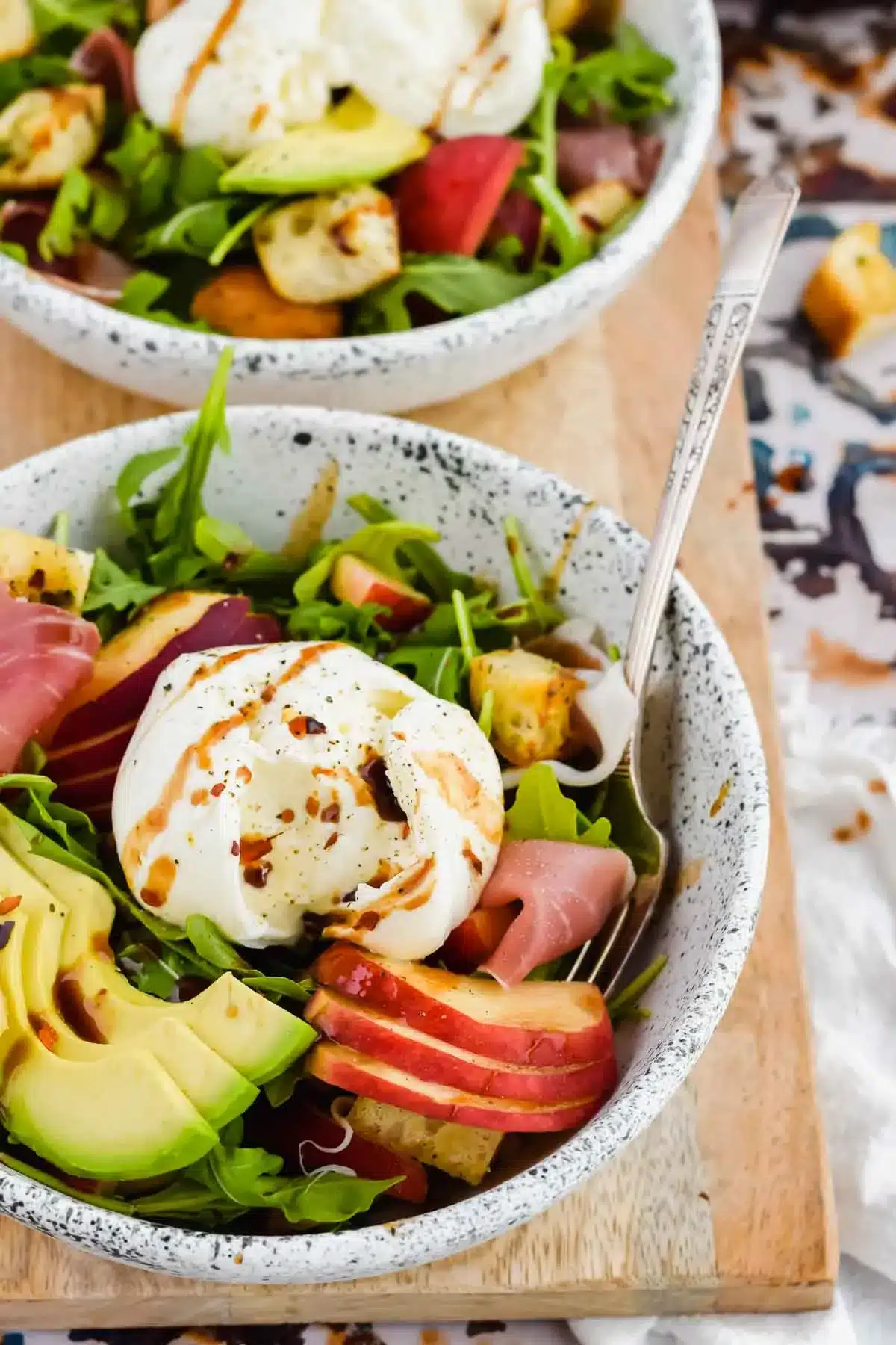 Peach burrata salad with avocado, and prosciutto in a white speckled bowl.