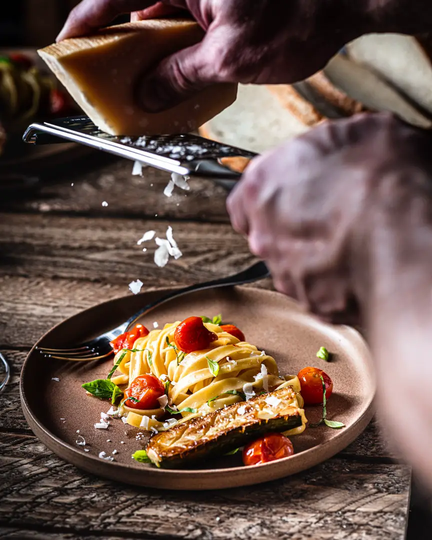 Hands grating cheese over pasta dish with blistered cherry tomatoes.