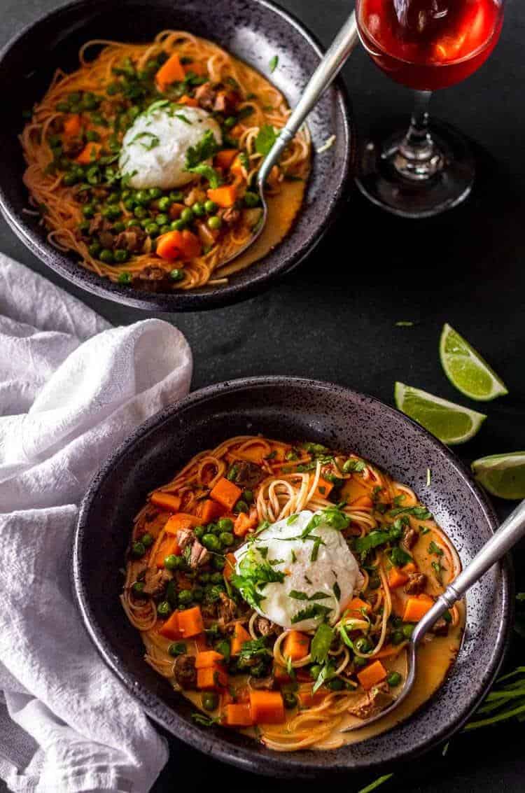 Peruvian beef noodle soup in a dark colored bowl with lime wedges in the background.