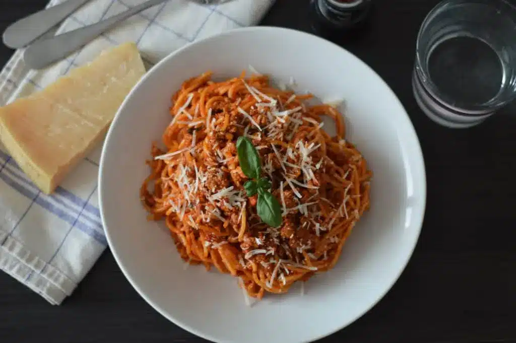 Spaghetti bolognese in a white bowl with a glass and cheese in the background.