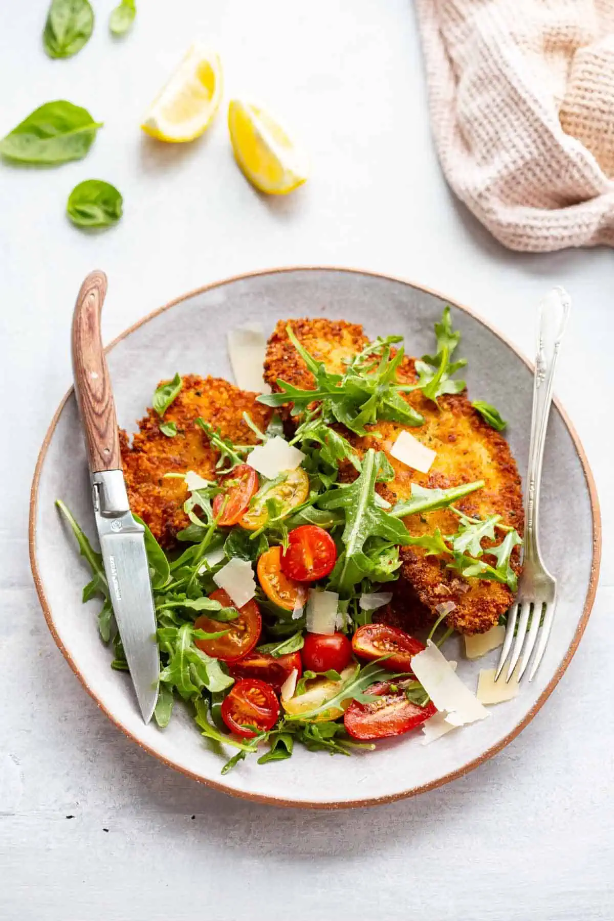 Crispy chicken Milanesa with tomato arugula salad on a white plate with fork and knife.