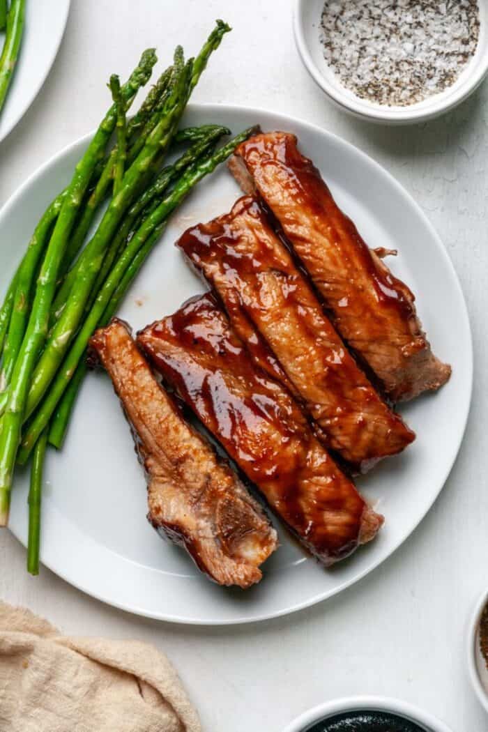 Barbecue covered ribs with asparagus on a white plate.