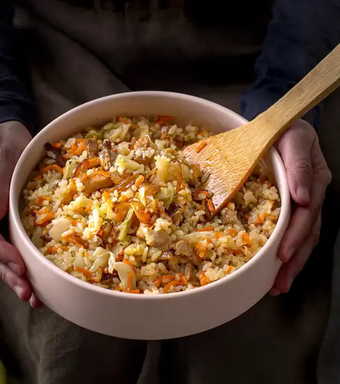 Hands holding bowl of rice with pork and veggies.