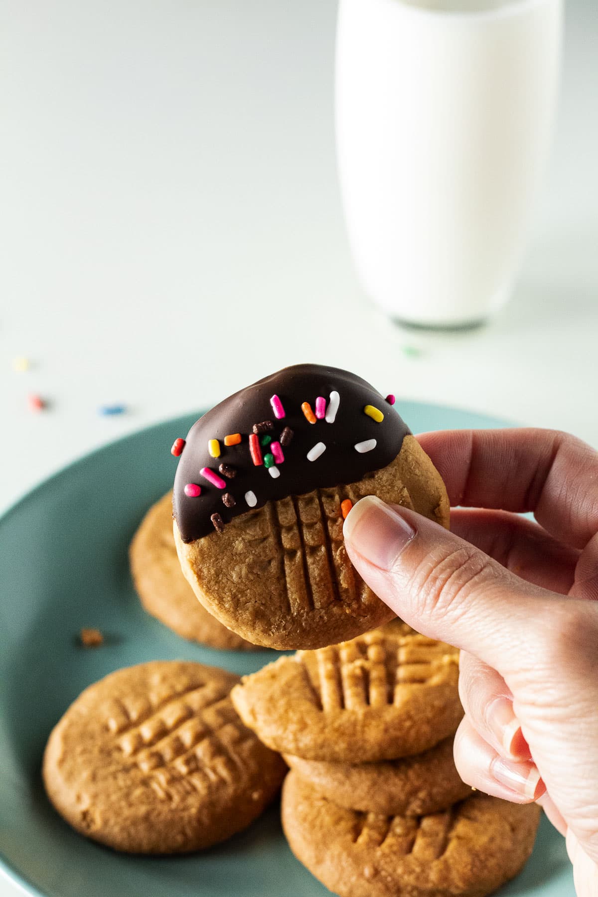 Hand holding chocolate dipped peanut butter cookie with sprinkles with a glass of milk in the background.