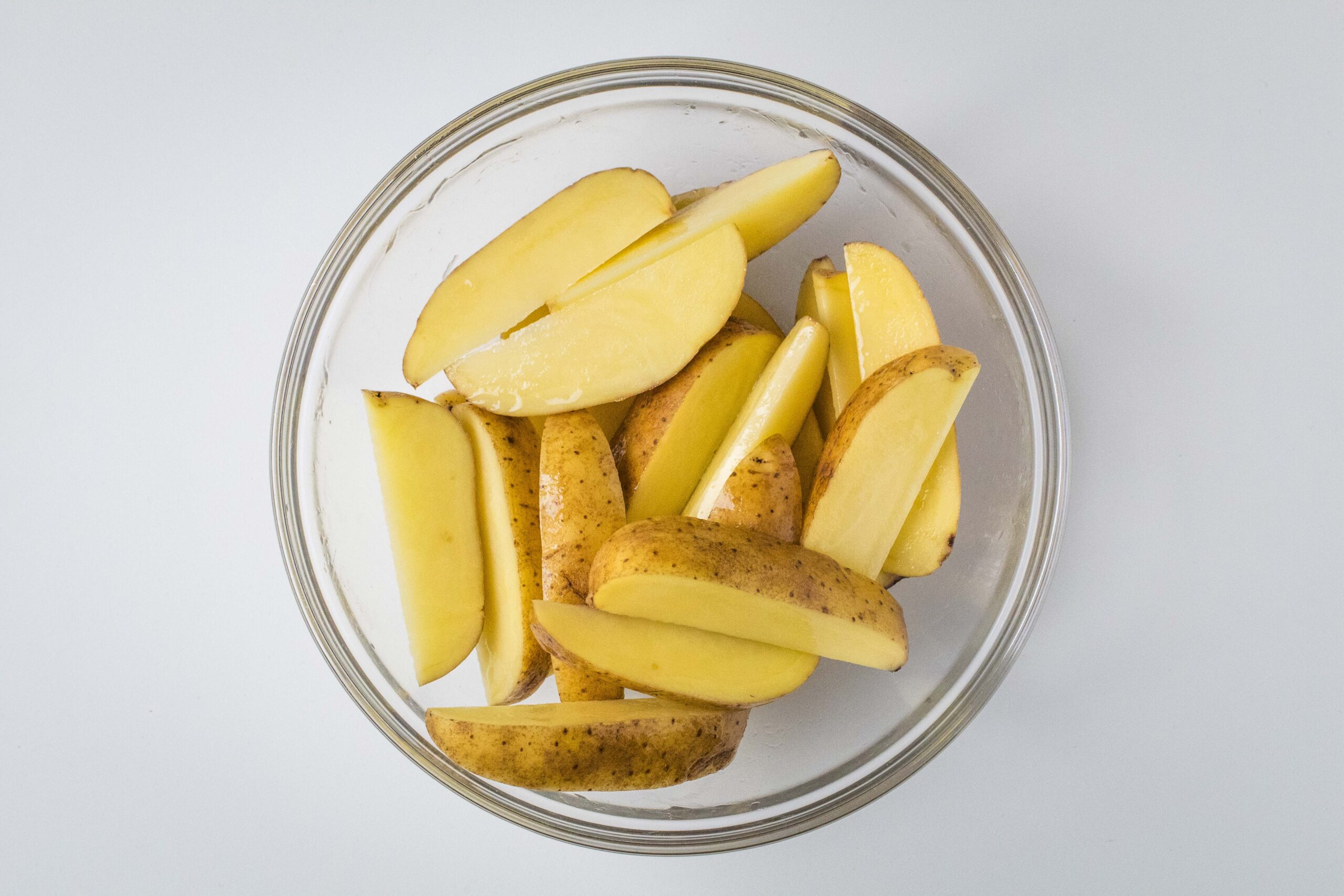 Raw potato wedges with oil in a glass bowl.