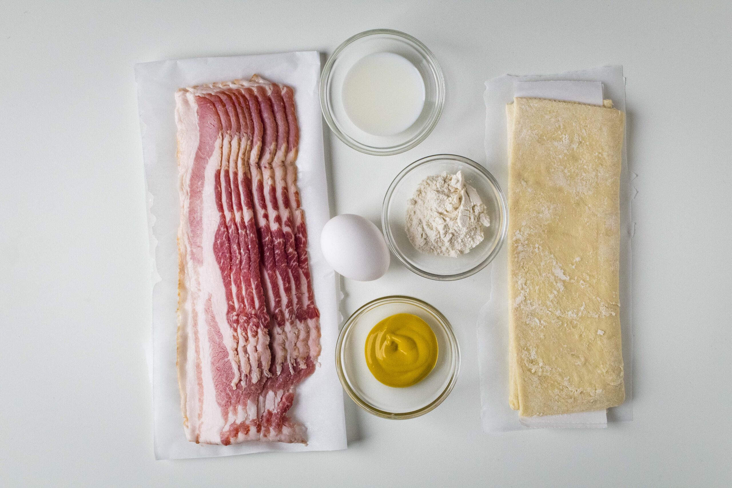 Ingredients for bacon twists including bacon, puff pastry, egg, mustard, flour, and milk.