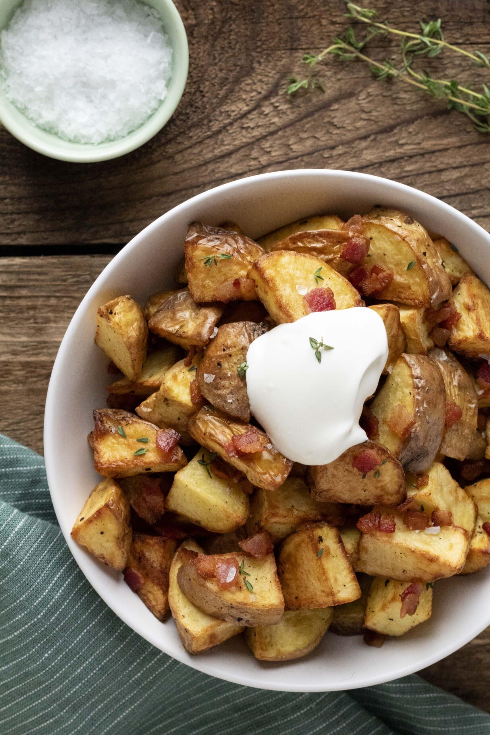 Air fryer crispy roasted potatoes with bacon, salt, herbs, and sour cream in a white bowl on a wooden background.