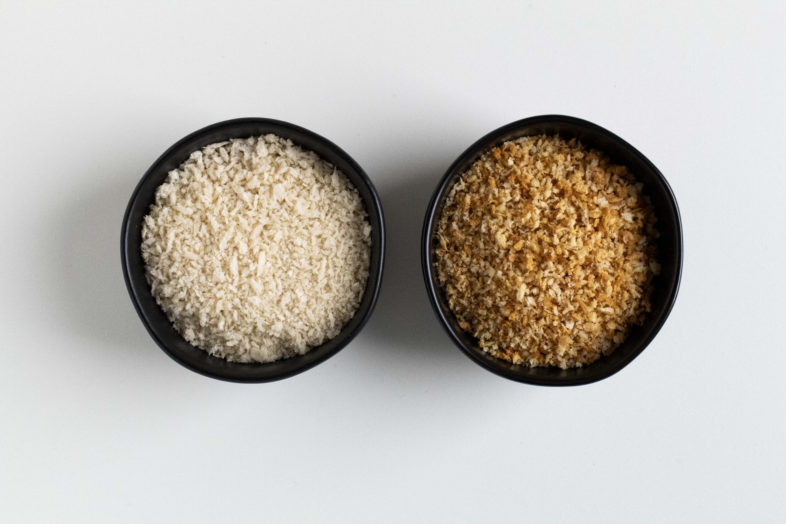 Untoasted and toasted breadcrumbs in black bowls side by side.