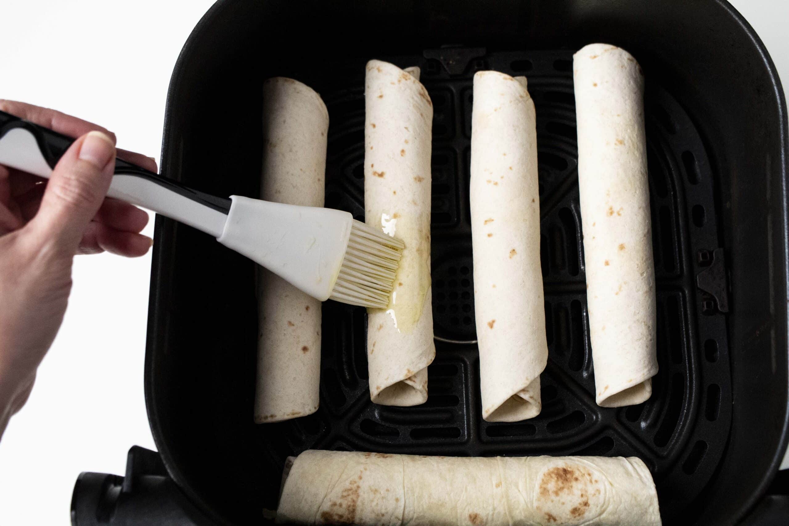 Olive oil brushed on taquitos inside an air fryer basket.