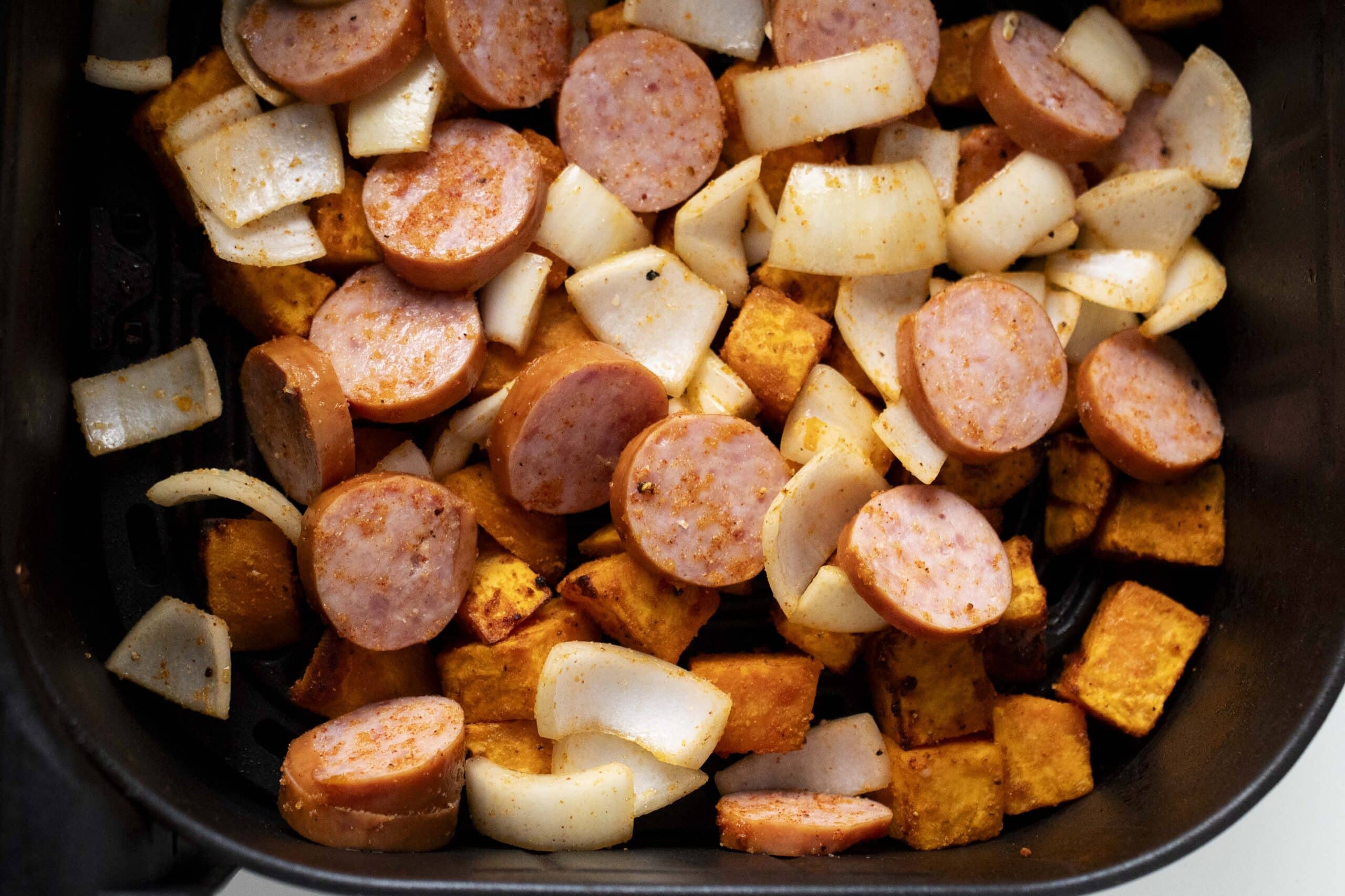 Diced sweet potatoes with sliced sausage and onions in an air fryer basket.