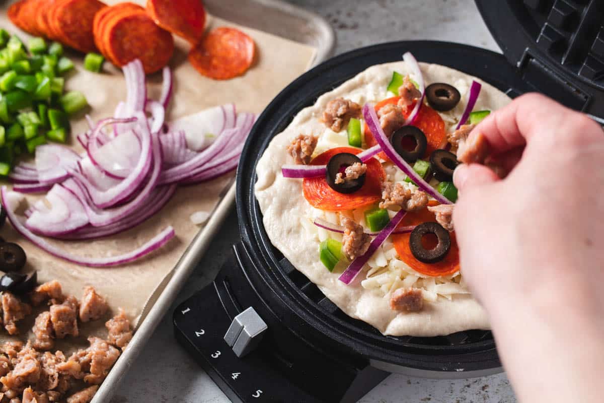 Adding toppings to pizza dough on waffle iron,