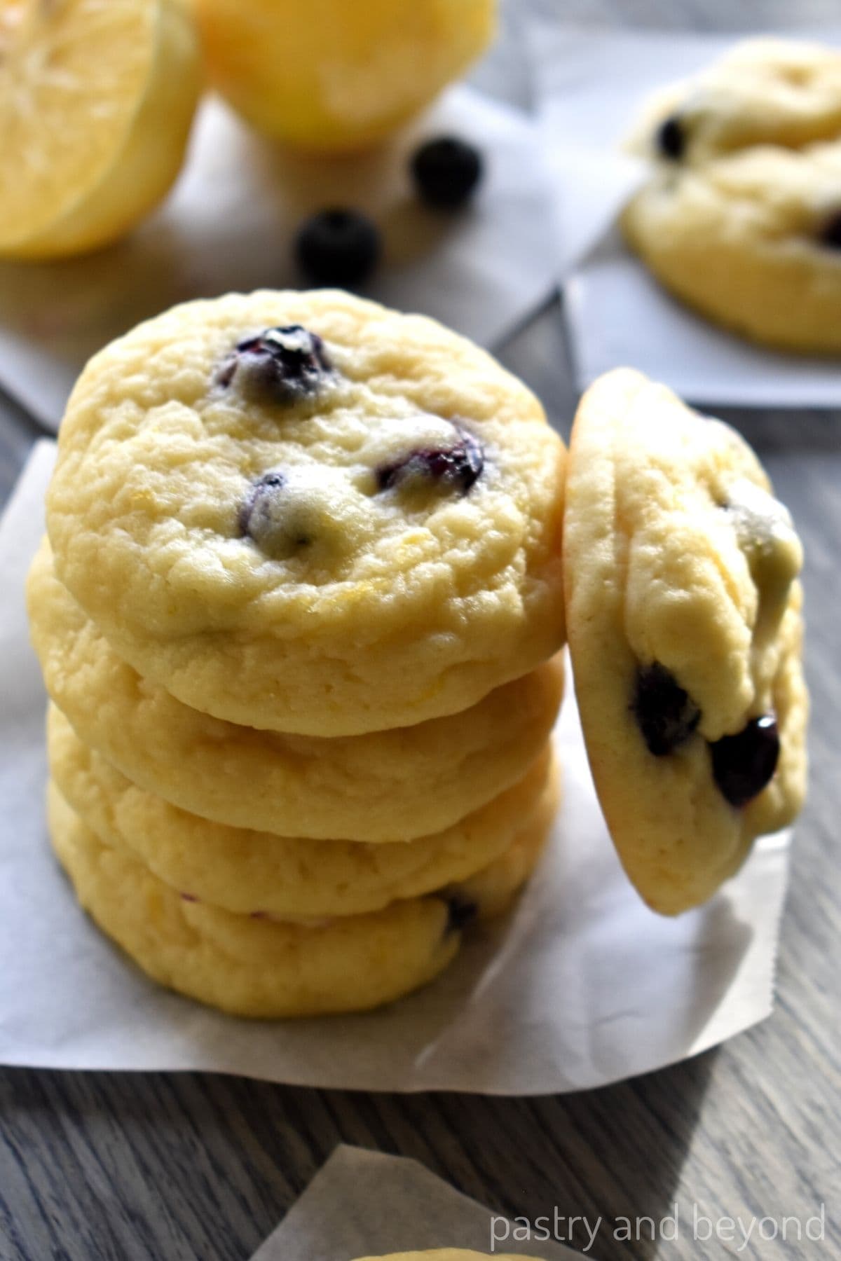 A stack of soft lemon cookies with fresh blueberries, placed on parchment paper with whole blueberries and lemon halves nearby.
