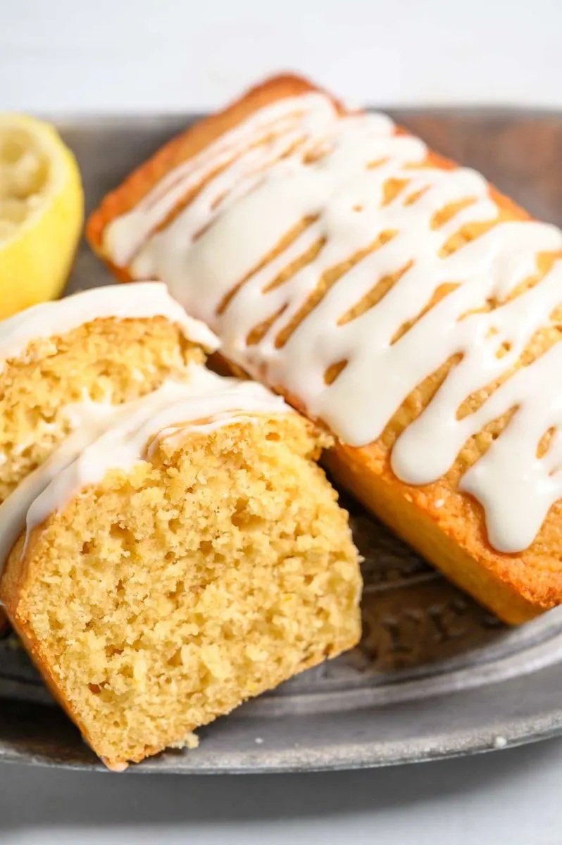 A lemon loaf drizzled with white icing, one loaf partially sliced, placed on a rustic tray with a lemon in the background.