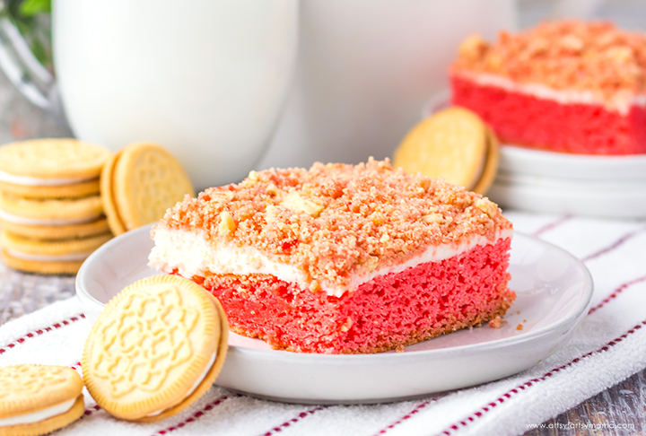 A slice of red crumb cake with a creamy topping and crushed cookie crumble on a white plate with cookies around.