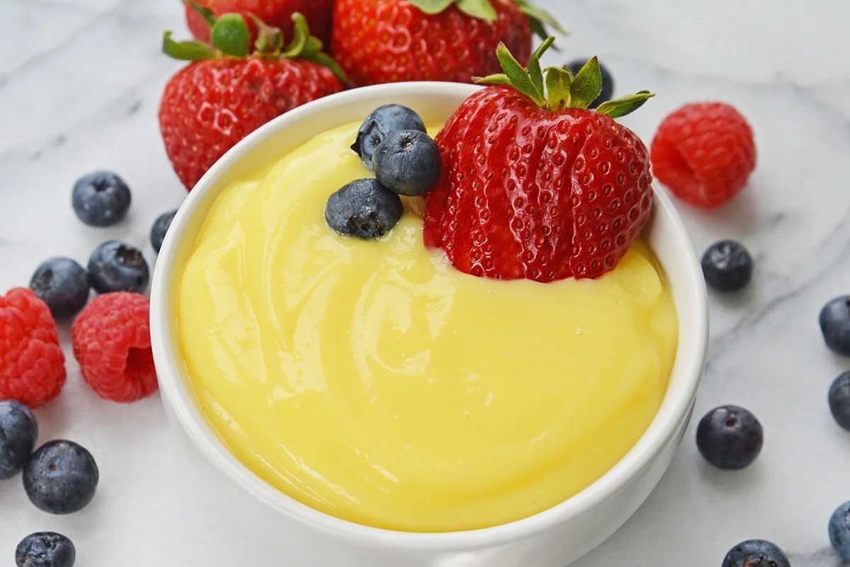 A bowl of vanilla custard topped with fresh strawberries, blueberries, and raspberries on a marble surface.