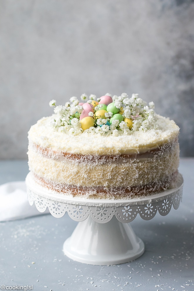 A layered coconut cheesecake cake on a white cake stand, decorated with shredded coconut, pastel chocolate eggs, and flowers.