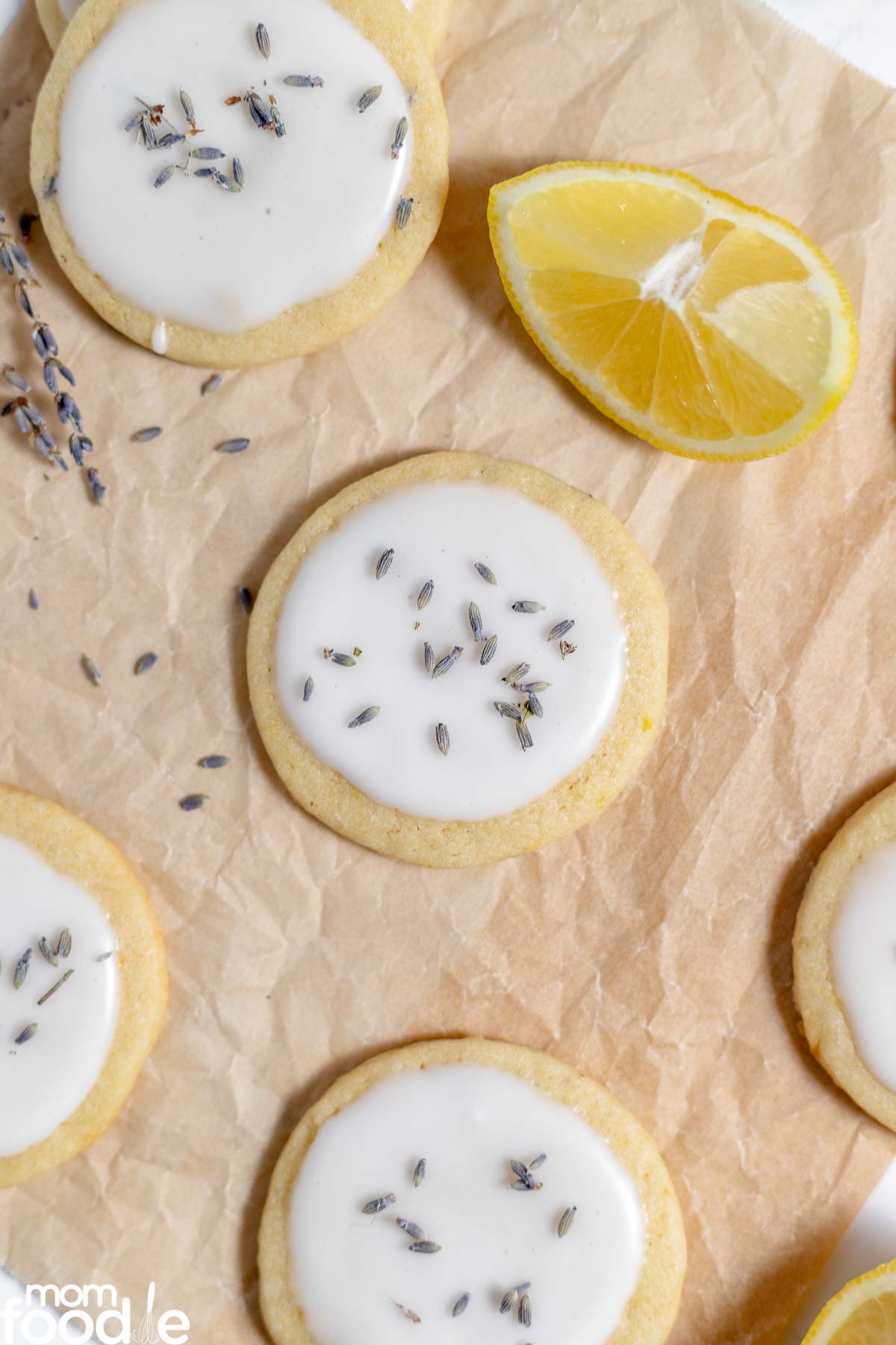 Round shortbread cookies with a white glaze, sprinkled with dried lavender, placed on parchment paper with a lemon wedge.