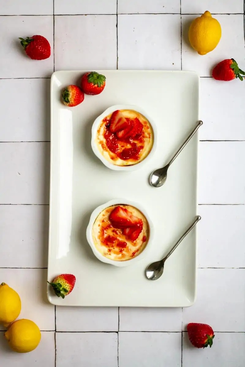 Two ramekins filled with of strawberry ricotta cheesecake, garnished with fresh strawberries, placed on a white tile background.