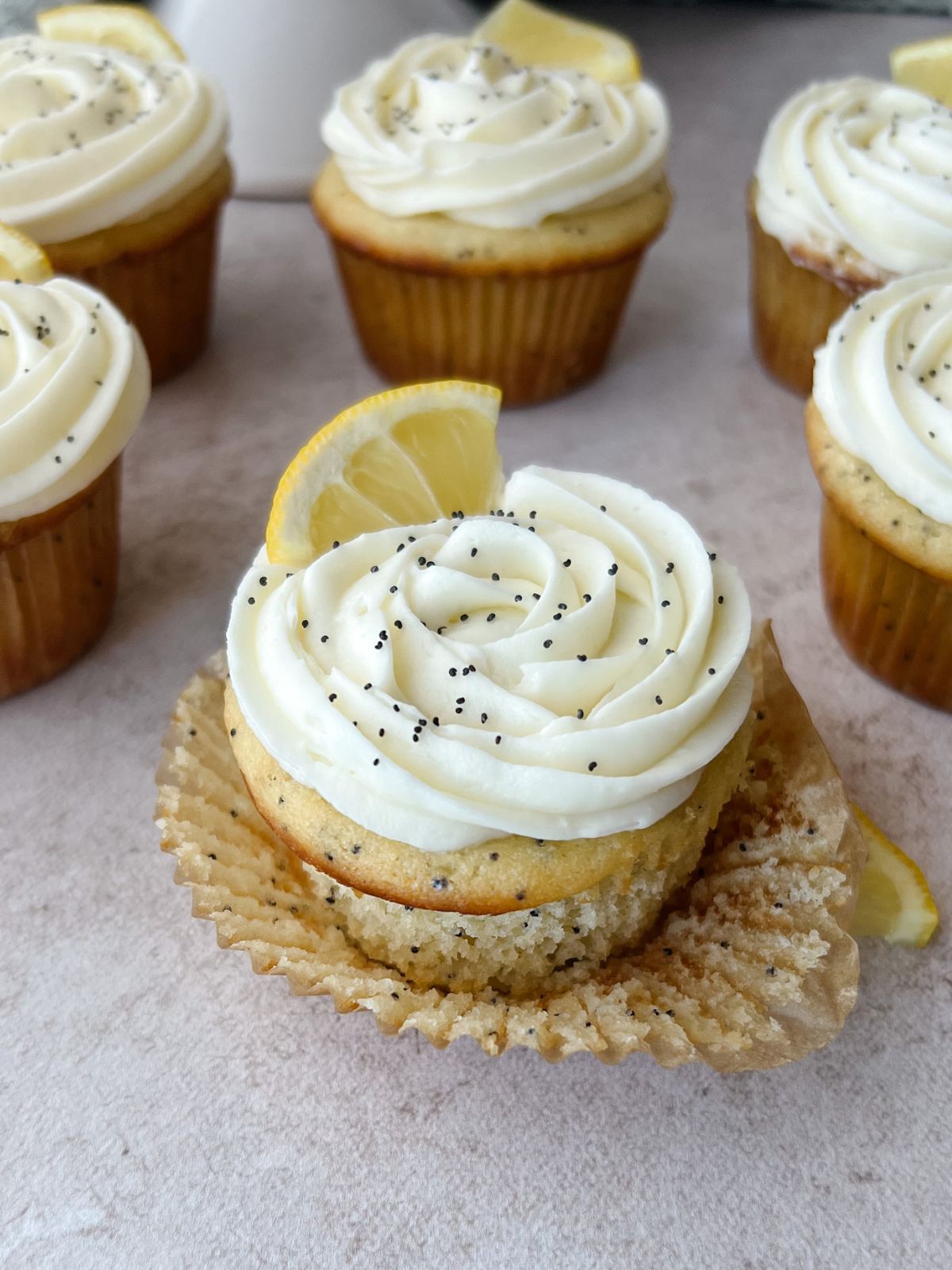 A cupcake with white frosting, lemon slice garnish, and poppy seeds on a beige surface.