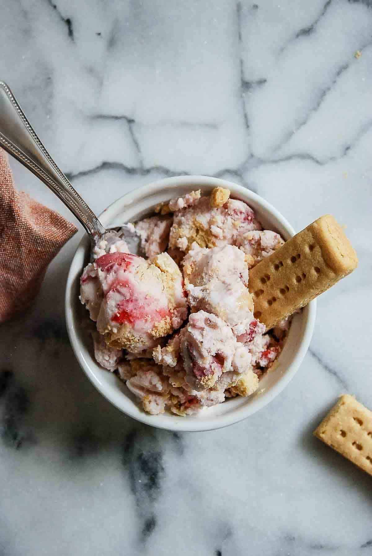 A bowl of strawberry shortcake ice cream with shortbread chunks and a biscuit stick on a marble surface.