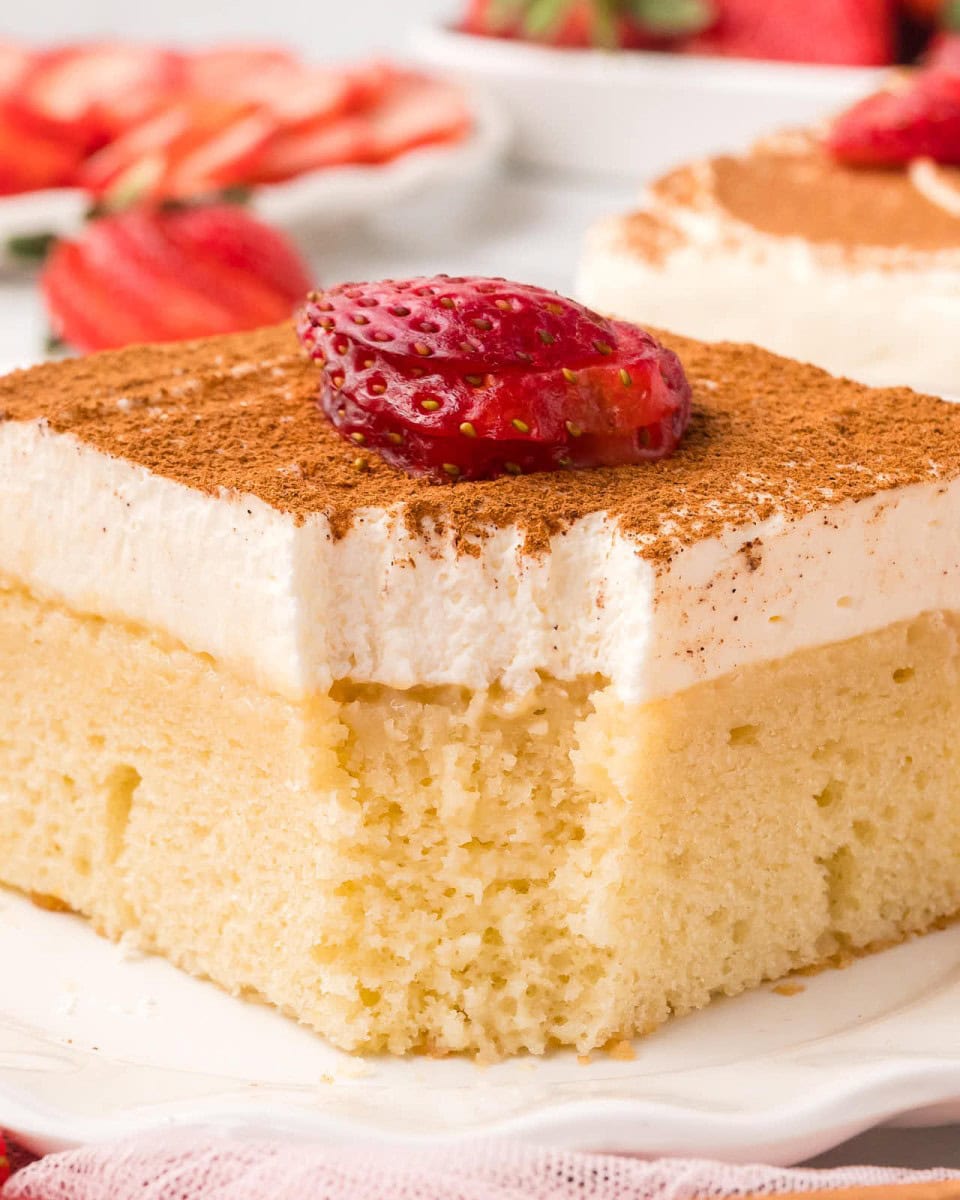 A square of tres leches cake with whipped topping, cinnamon, and a strawberry slice on a white plate.