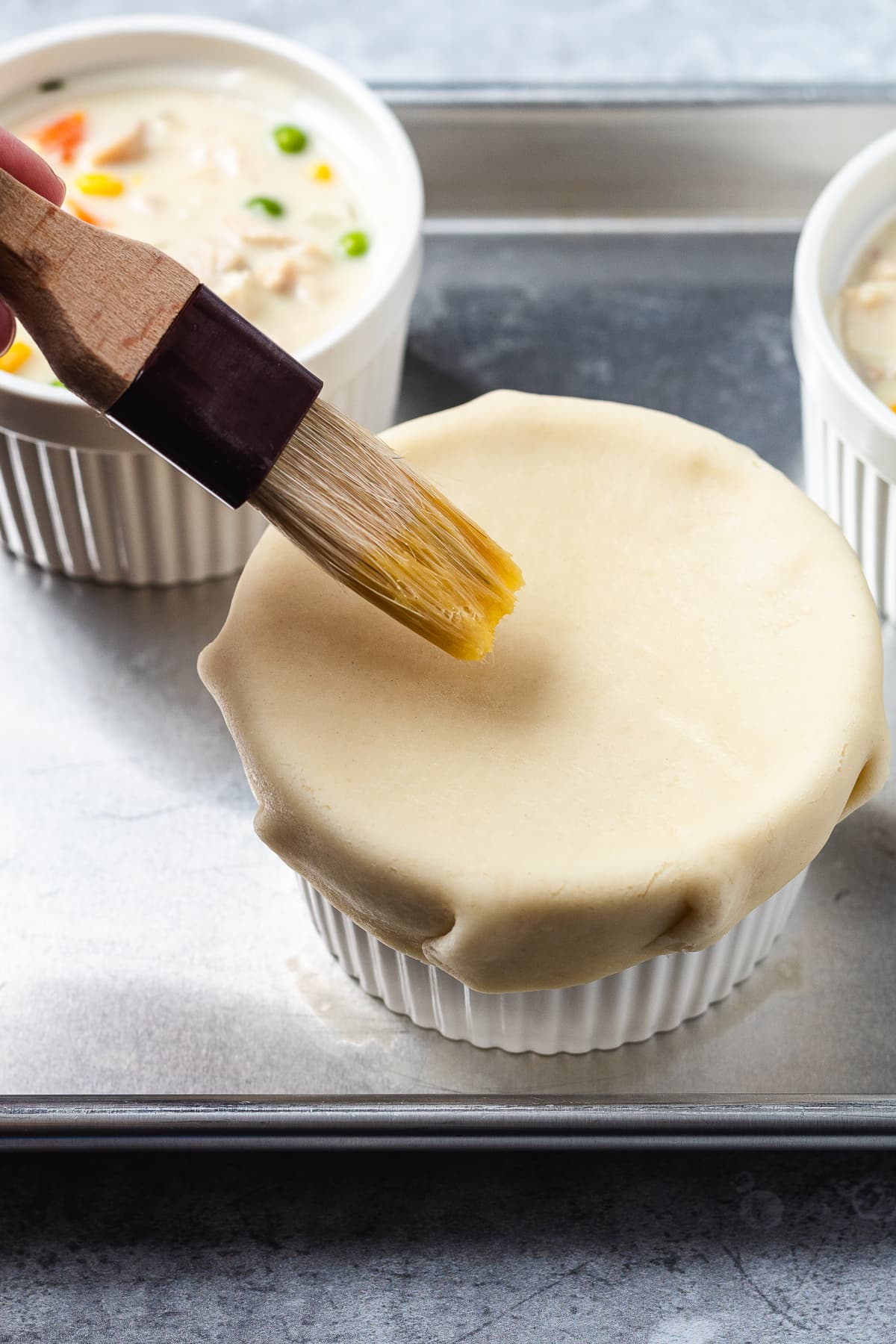 Brushing egg wash over pie crust on a mini chicken pot pie in a white ramekin.