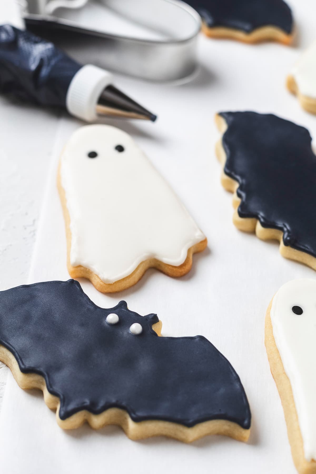 Iced Halloween bat and ghost cookies arranged on a white surface with piping bag and cutter nearby.