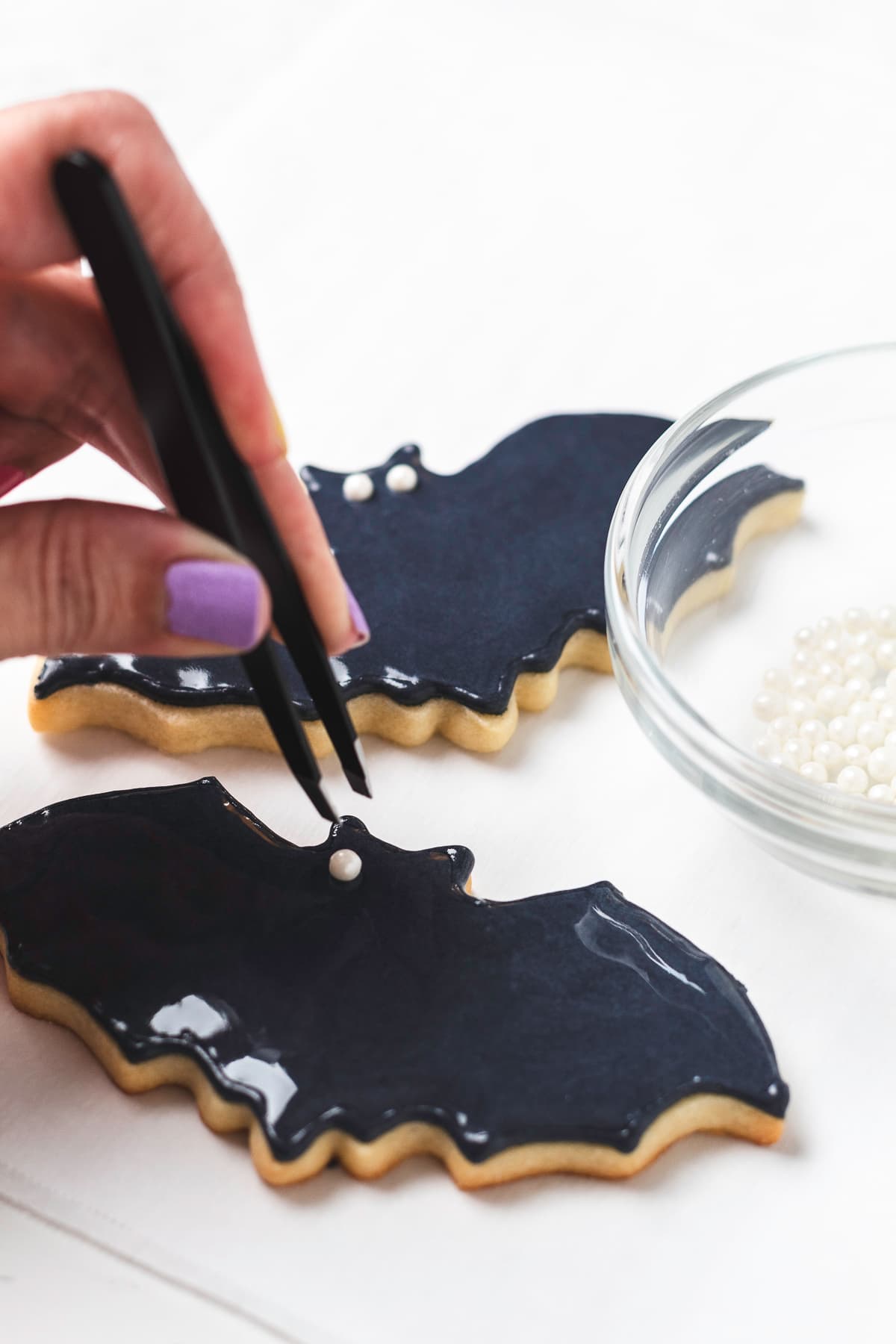 Tweezers placing sugar pearl eyes onto a bat-shaped sugar cookie with black icing.