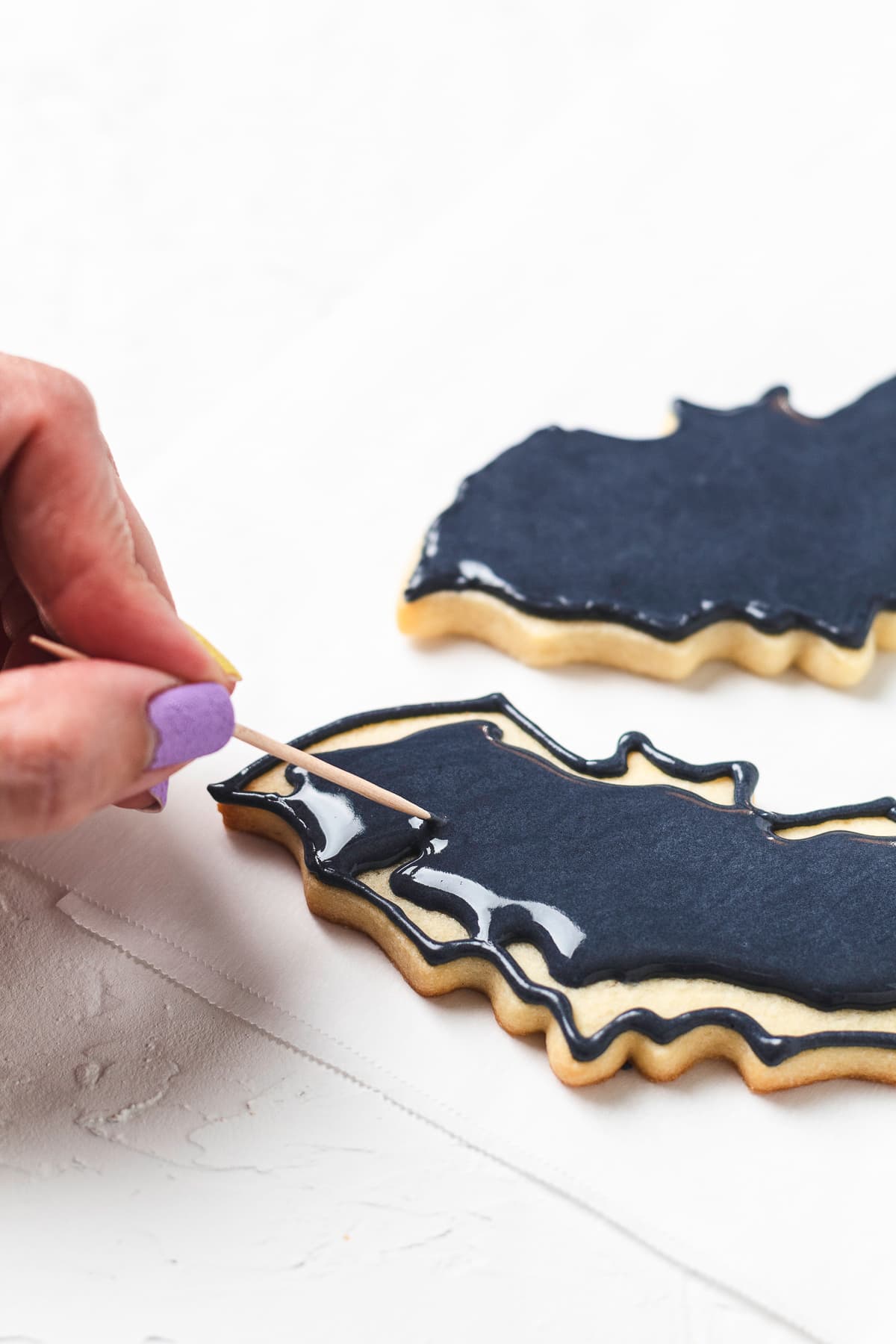 A toothpick spreads black icing across a bat-shaped sugar cookie to smooth the surface.