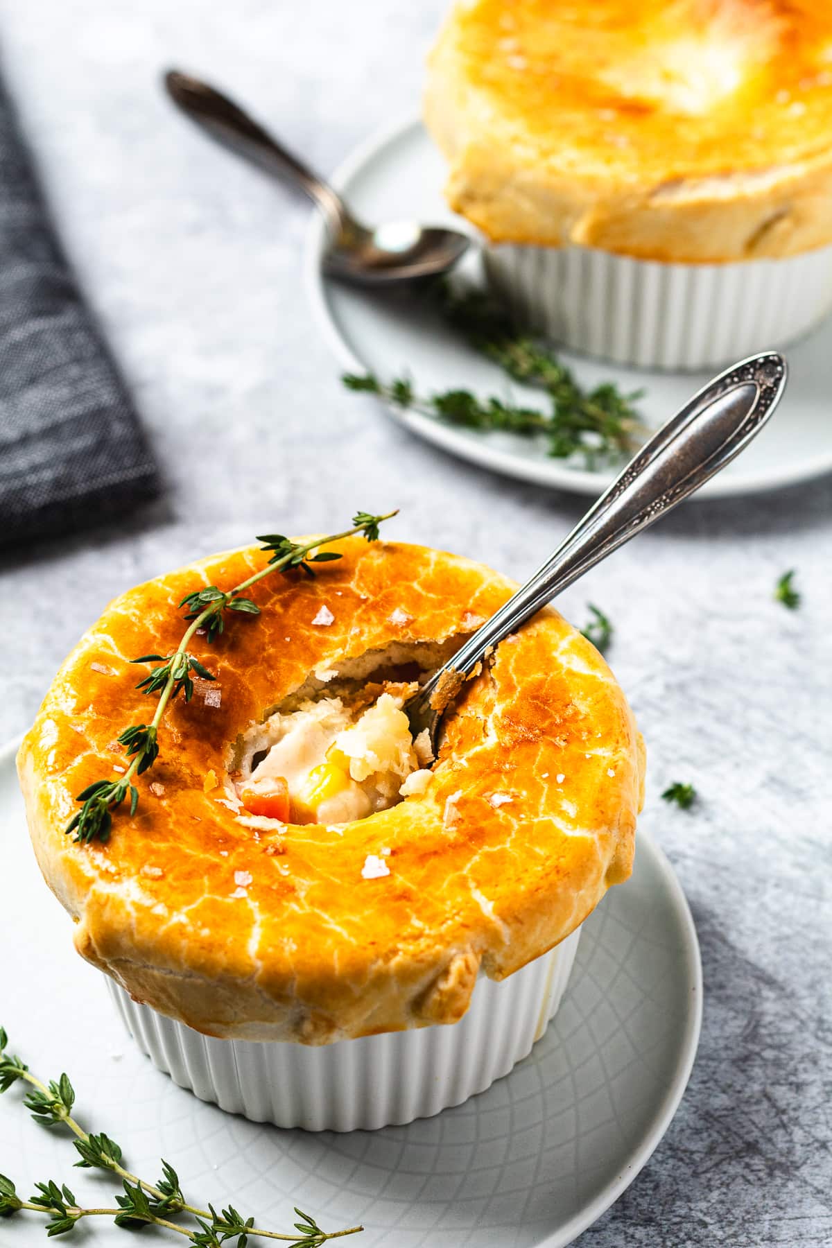 Golden brown baked chicken pot pie in a ramekin with a spoon and thyme garnish on a white plate.
